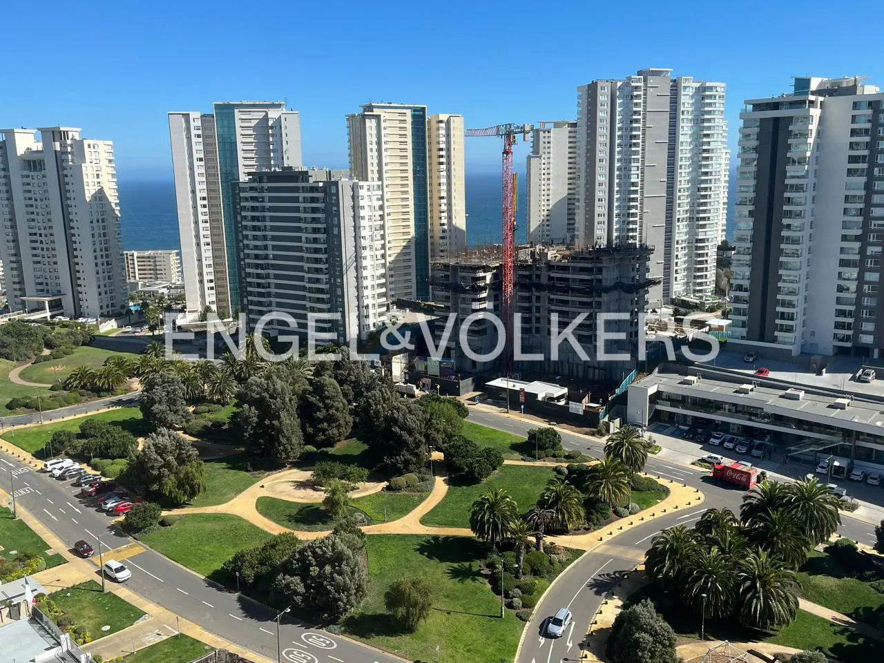 Cityscape view with tall buildings, a park with green trees, and a construction site with a red crane under a blue sky.