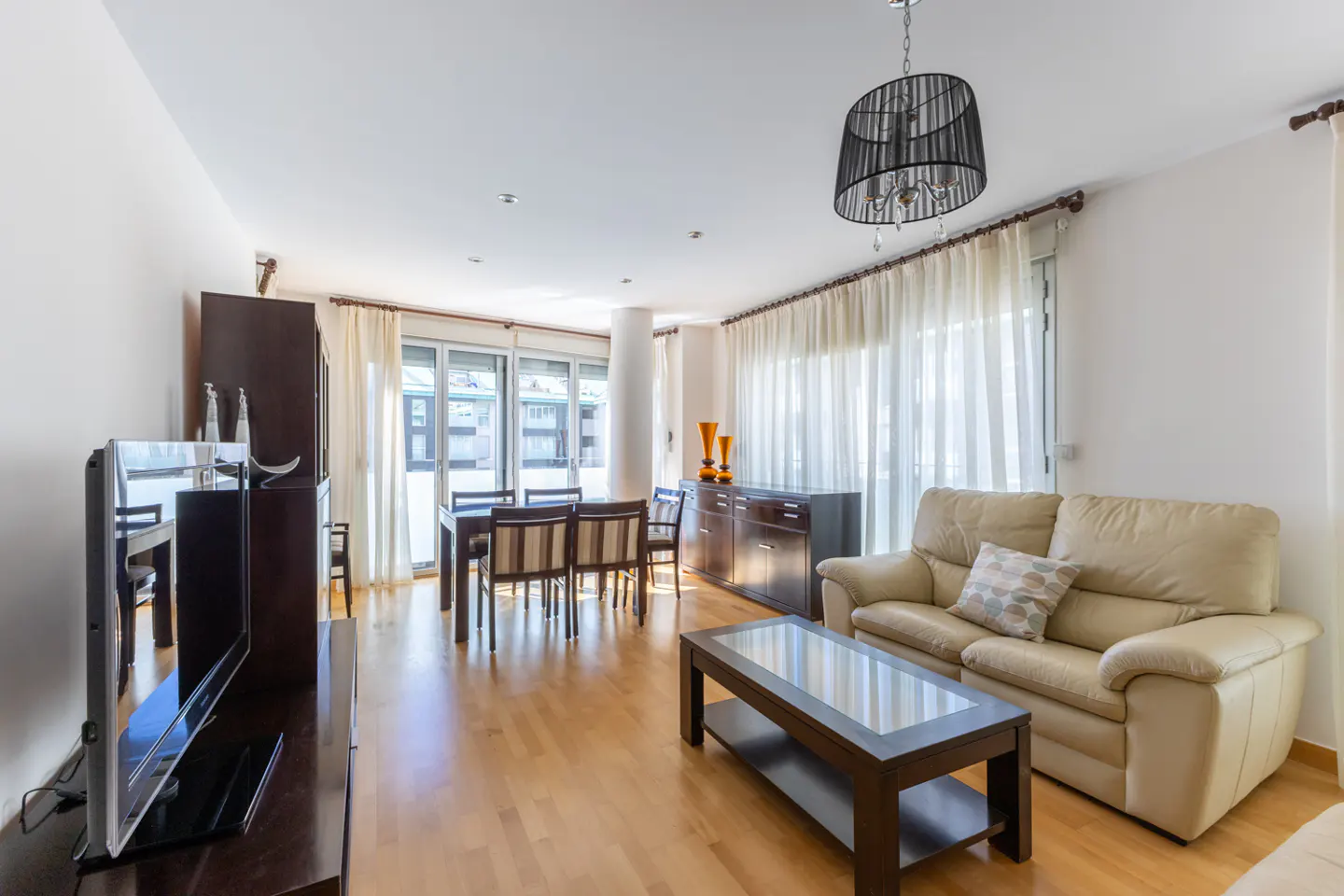 Bright living room with wood floors, a beige sofa, a dark wood coffee table, and a dining table near the window.