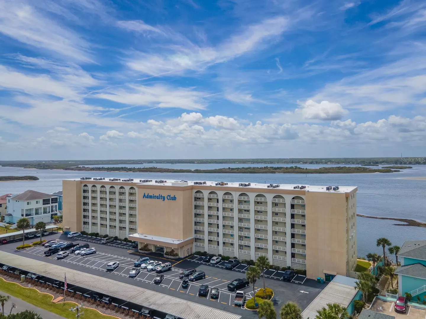 Aerial view of the Admiralty Club, a beige condo building with a parking lot, near a body of water under a blue sky.