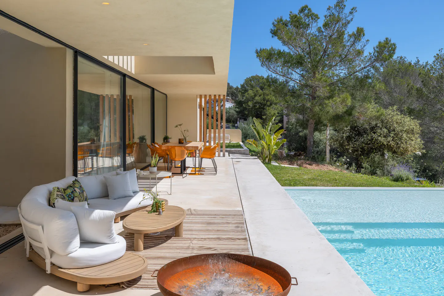 Outdoor patio with white sofa, wood tables, and rusty fire pit. A pool and green trees are in the background.
