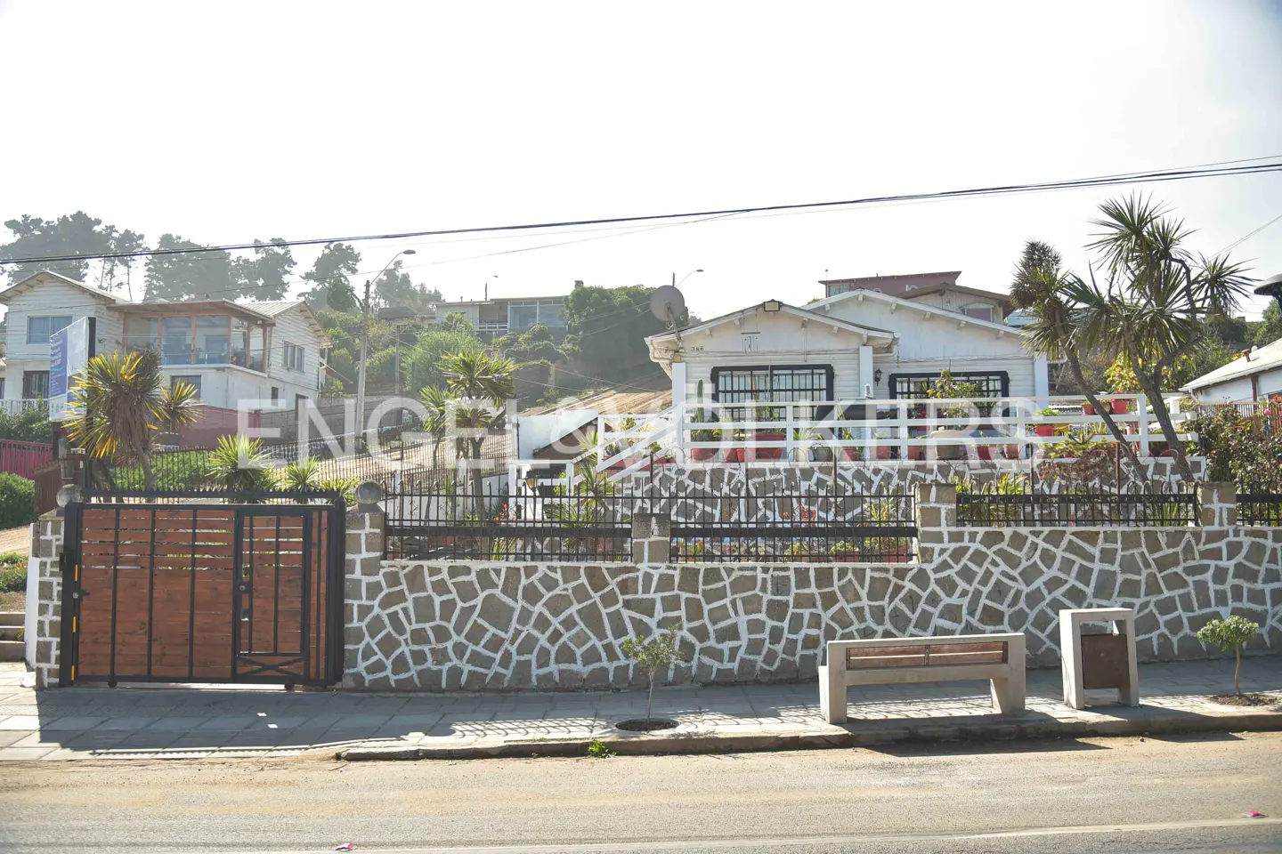 Exterior view of a white house with a stone wall and a wooden gate. A bench and trash can sit on the sidewalk.
