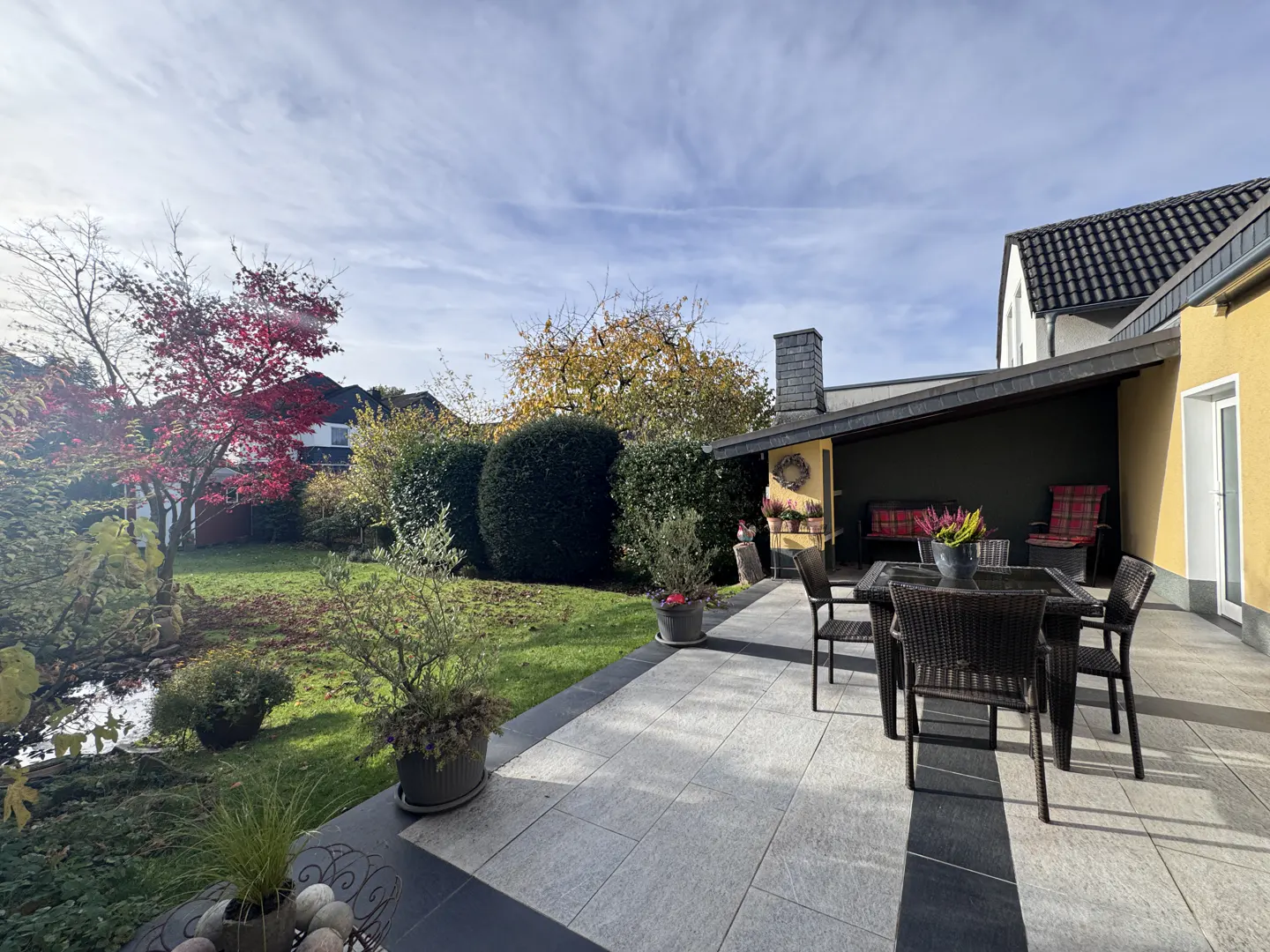 A paved patio with a dining set overlooks a lush backyard garden with a small pond and vibrant autumn foliage.