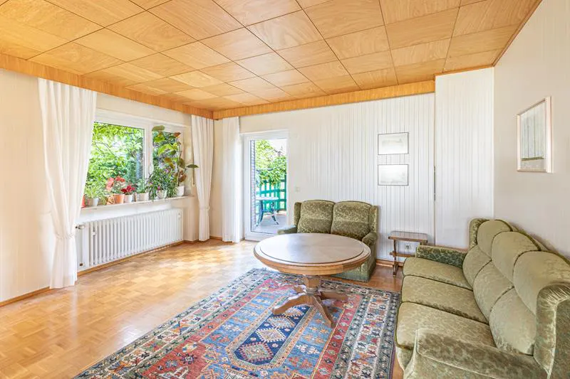 A living room with wood floors, a wood ceiling, and a colorful rug. There is a green sofa and chair, a round table, and a window with plants.