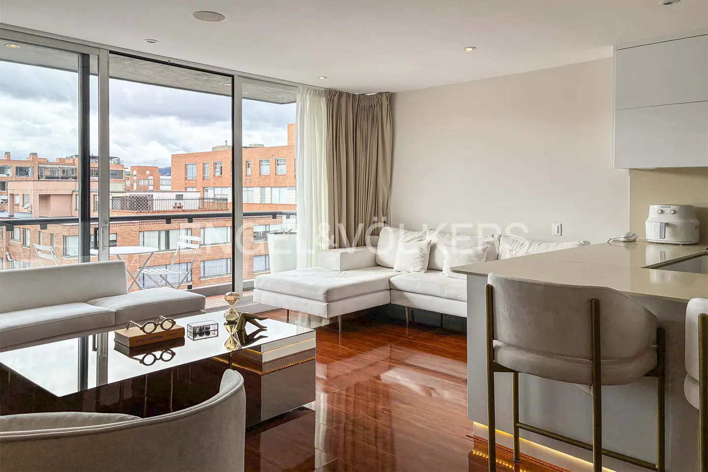 Bright living room with white sectional sofa, glass coffee table, and wood floors. Large windows overlook city buildings.