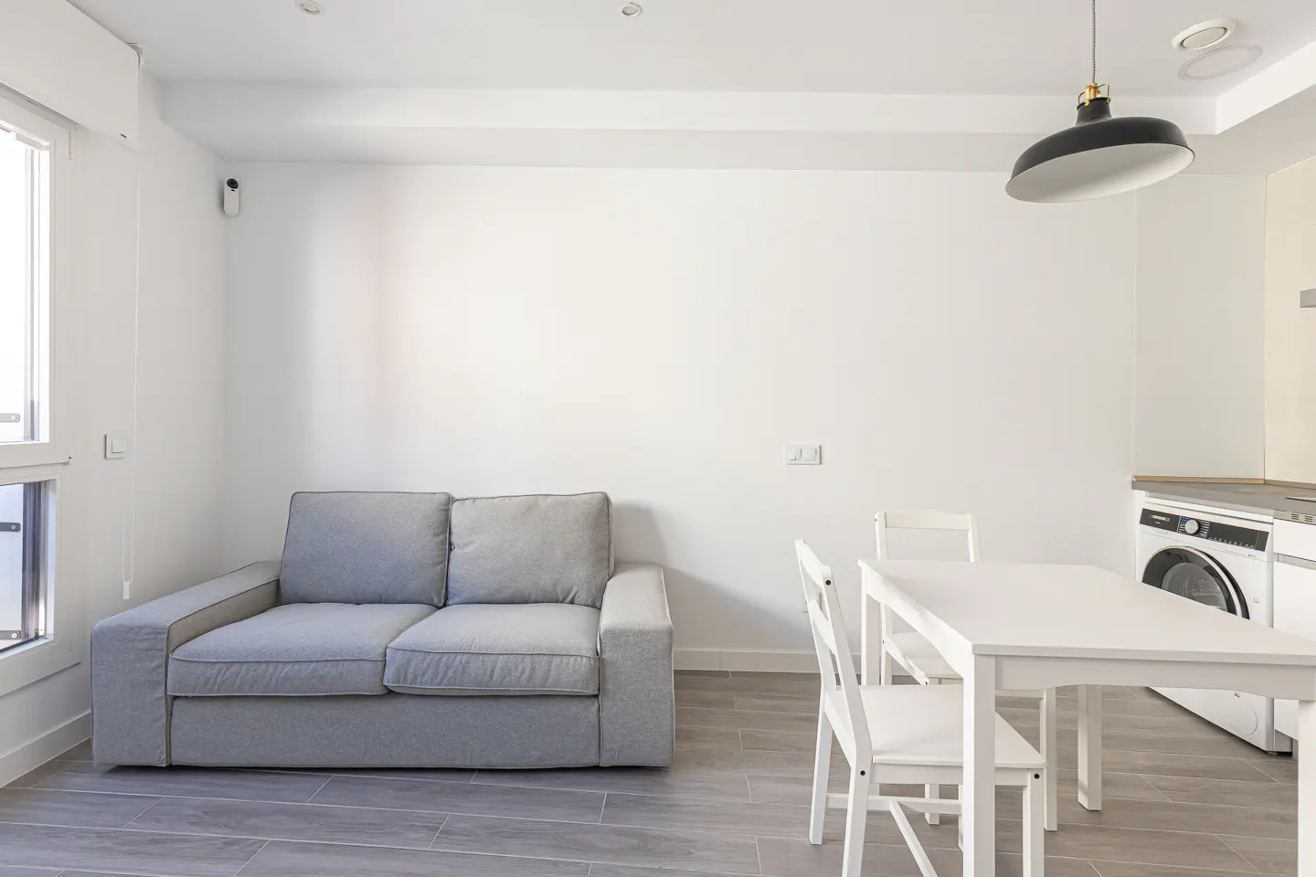 Modern studio apartment with a grey sofa, white dining table and chairs, and a built-in washing machine.