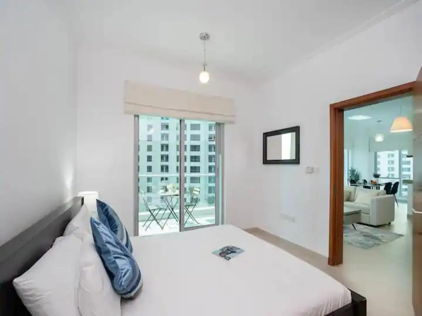Modern bedroom with white walls, a bed with blue pillows, a balcony view, and a doorway leading to a bright living area.