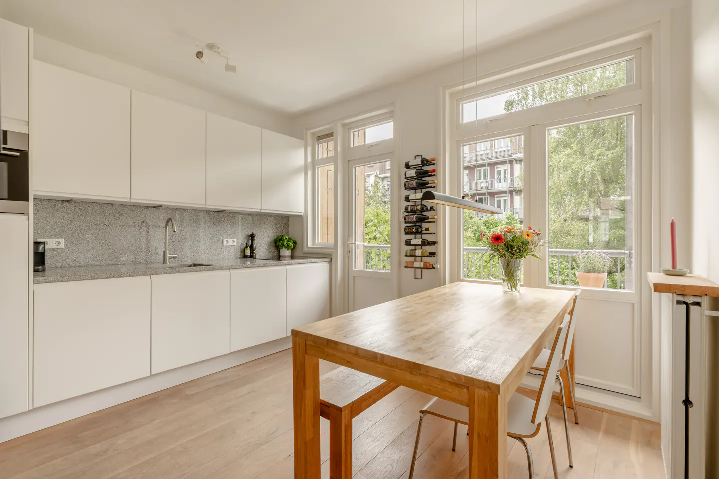 Modern kitchen with white cabinets, granite countertops, wooden dining table, and a wall-mounted wine rack by the balcony.