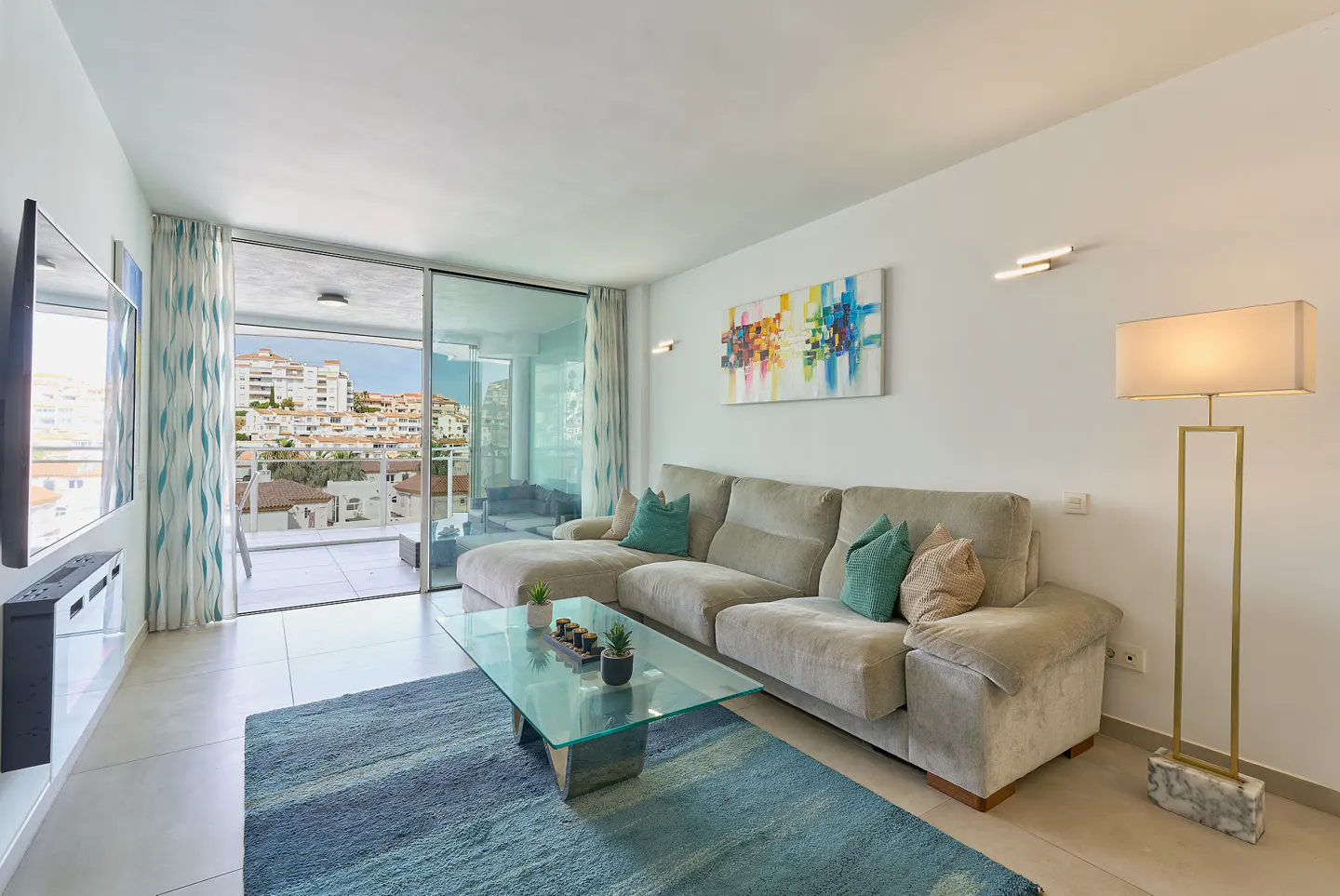 Bright living room with a beige sectional sofa, blue rug, glass table, and a view of a coastal town through sliding glass doors.