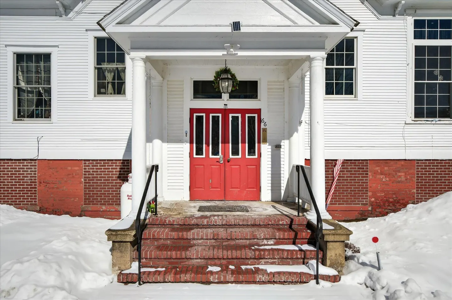 Exterior of a white building with a red door and brick steps covered in snow.
