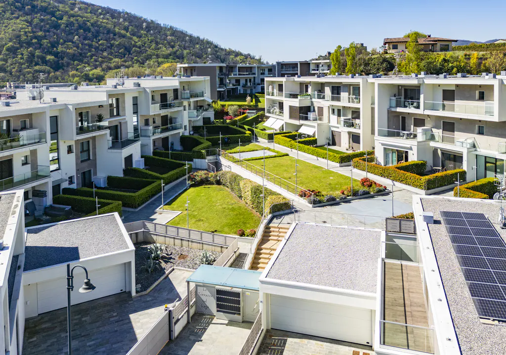 Aerial view of modern white townhouses with green lawns and trimmed hedges on a sunny day. Solar panels are visible on one roof.