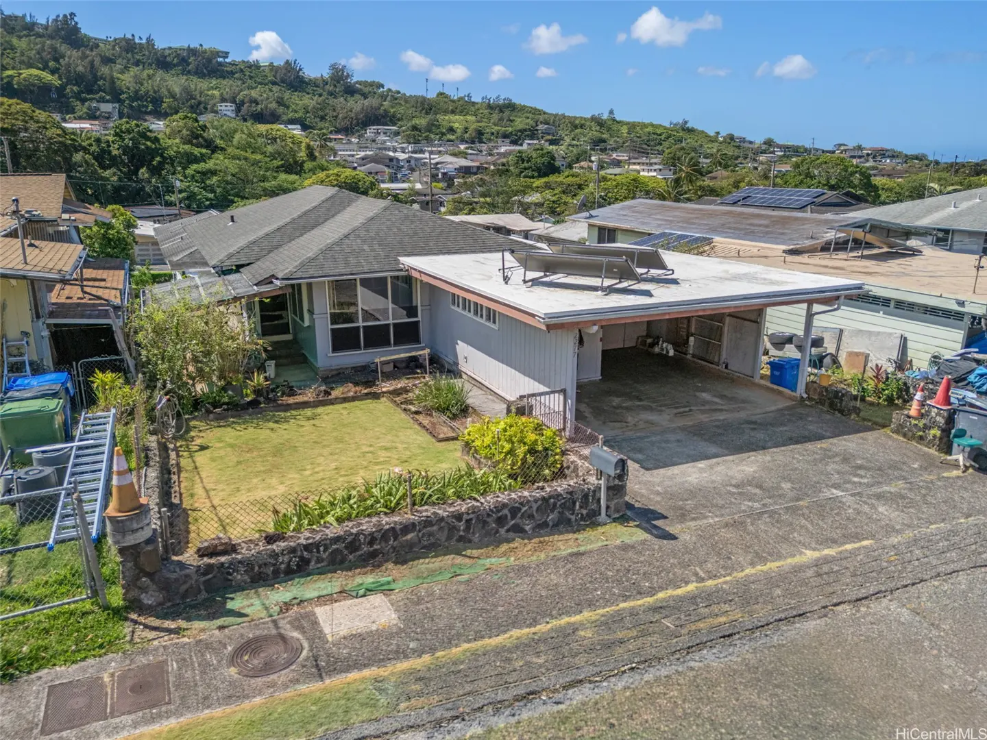 A single-story gray house with a carport and a green lawn in front of a lush, green hillside.