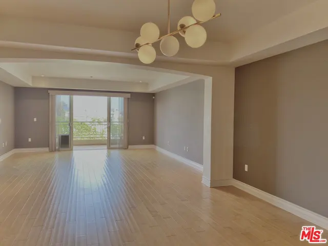 Empty living room with light wood floors, gray walls, and a modern chandelier. Sliding glass doors lead to a balcony with a view.