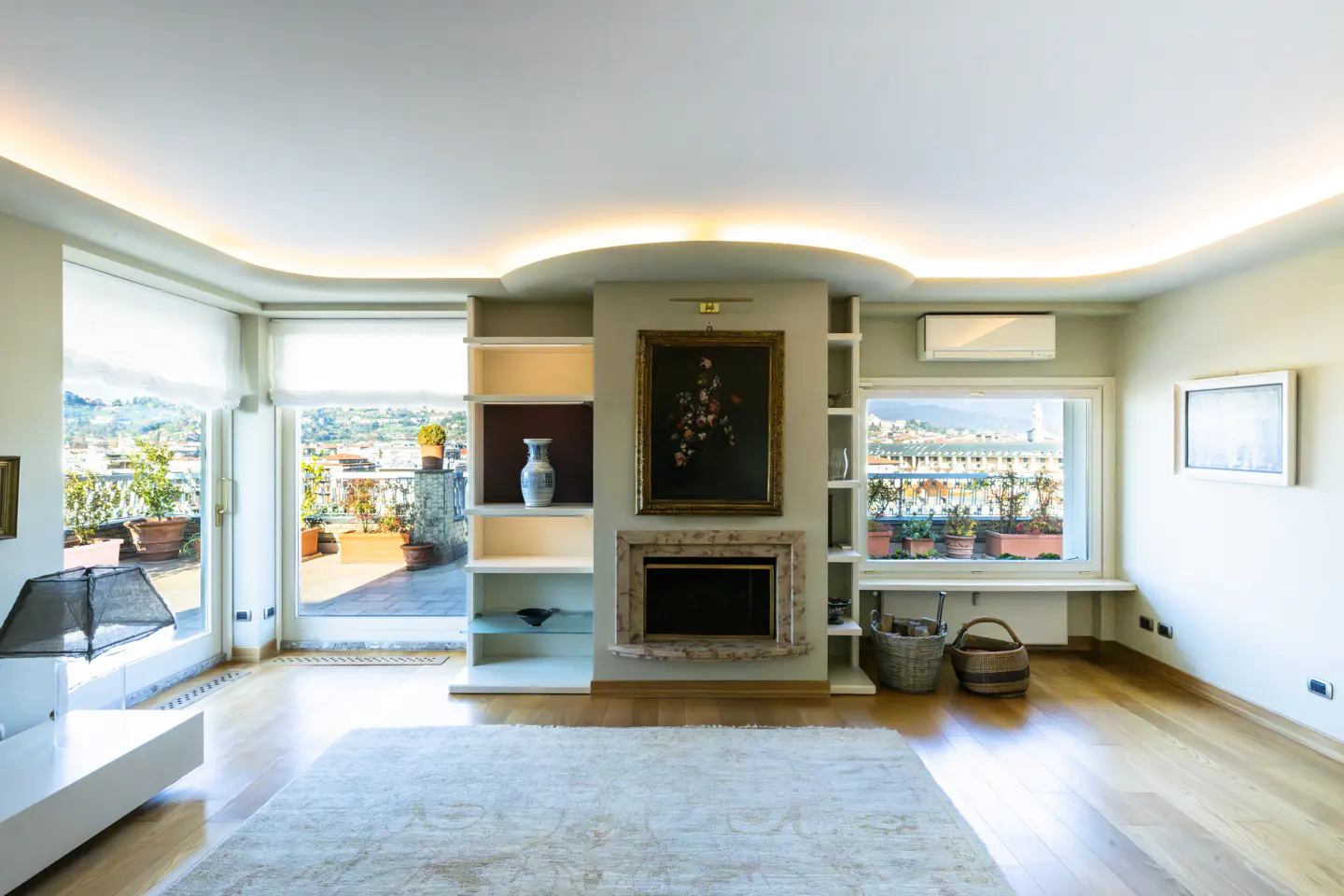 Bright living room with hardwood floors, a fireplace with a painting above, and built-in shelves. A large window shows a terrace with plants.