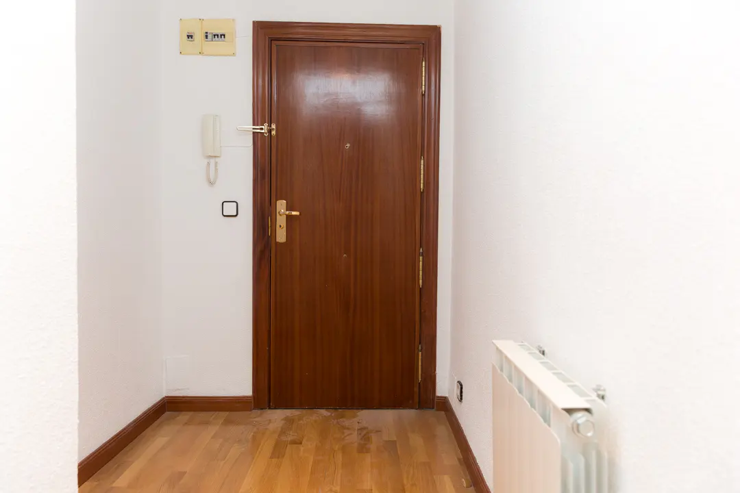 Entryway with a brown wooden door, light wood floors, and white walls. A white radiator is on the right wall.