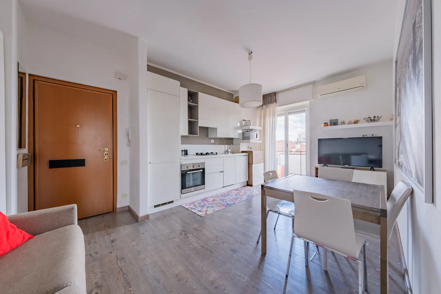 A bright apartment interior with a kitchen, dining table, and TV. White cabinets and walls contrast with the gray wood floor. A brown door is on the left.