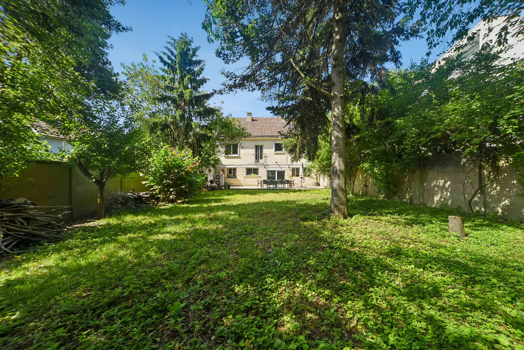 A large green lawn with trees surrounds a two-story beige house with a brown roof. A patio with a table and chairs is visible.