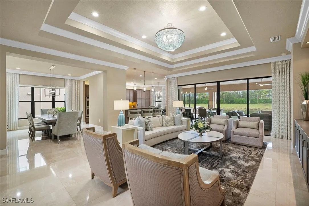 Open-concept living room with beige walls, marble floors, and a coffered ceiling with a crystal chandelier. Neutral furniture and a dining area are visible.