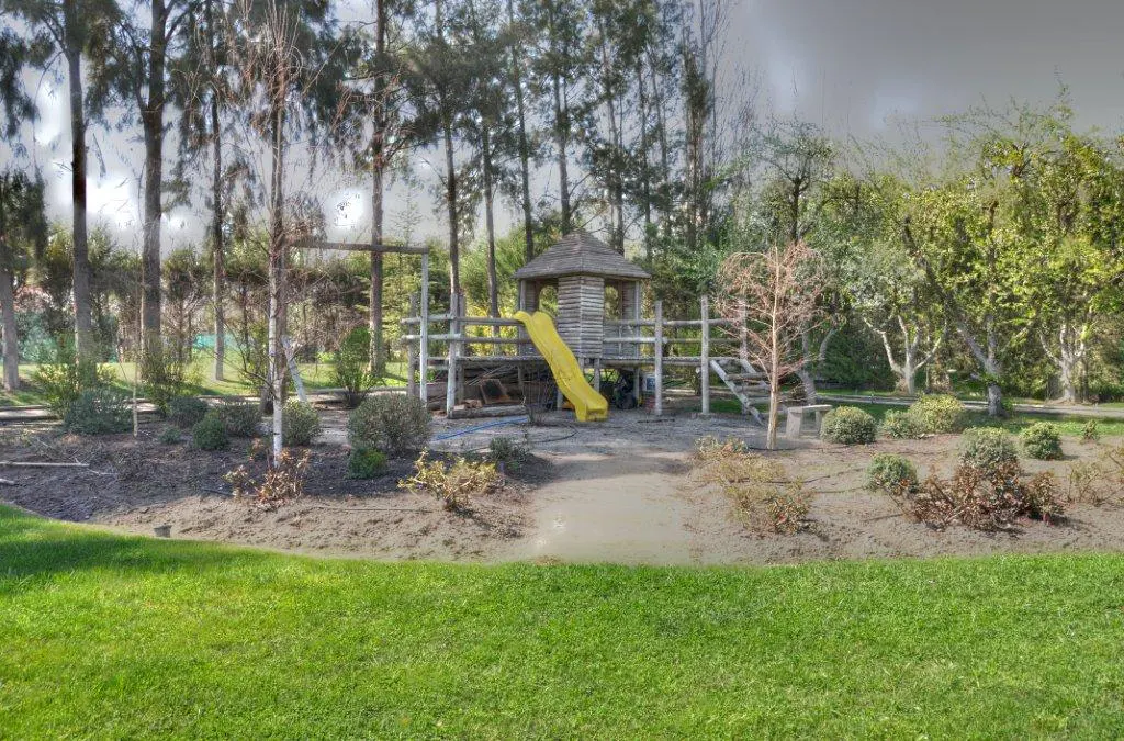 A wooden playground with a yellow slide sits in a yard with green grass and trees.