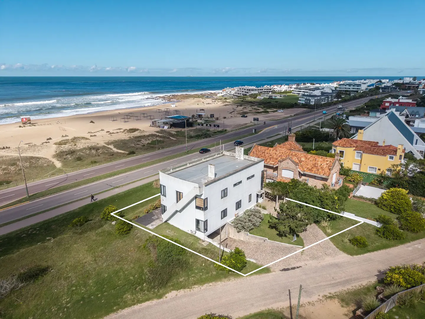 Aerial view of a modern white house with a white outline, near a beach and road on a sunny day.