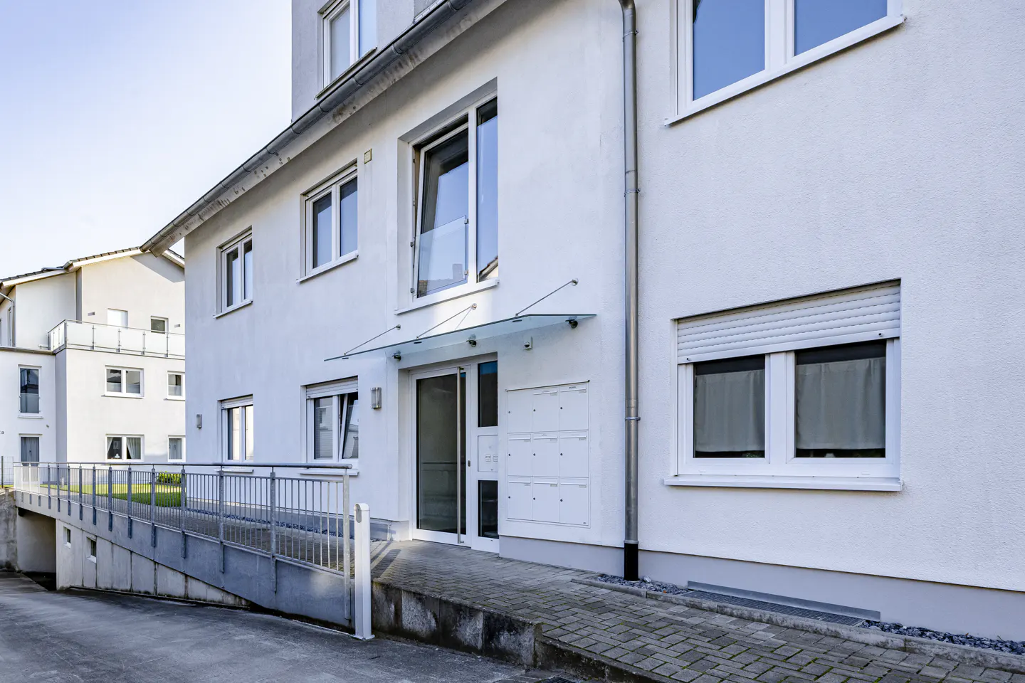 Exterior of a modern, white apartment building with a glass awning over the entrance and a ramp for accessibility.