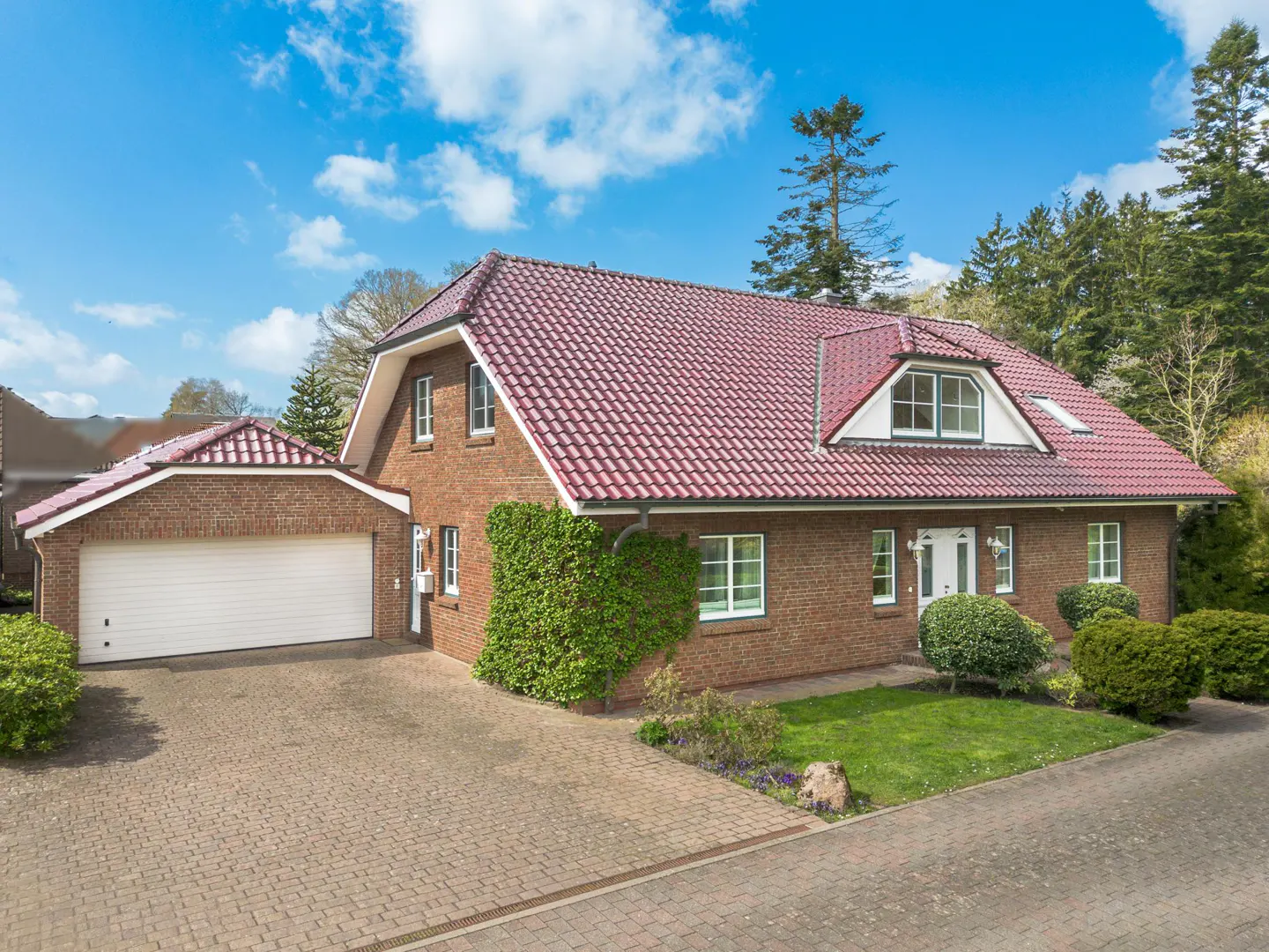 Brick house with a red tile roof, white trim, and a matching garage on a paved driveway. Green lawn and trees surround the property.