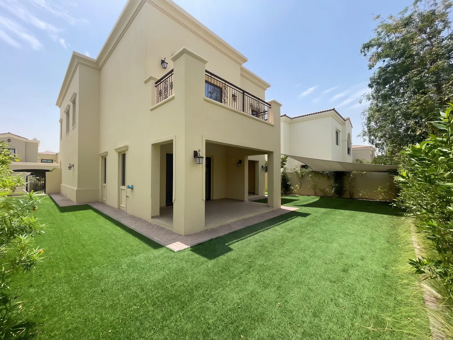 Two-story beige house with a balcony and a covered patio, surrounded by green grass and trees under a blue sky.