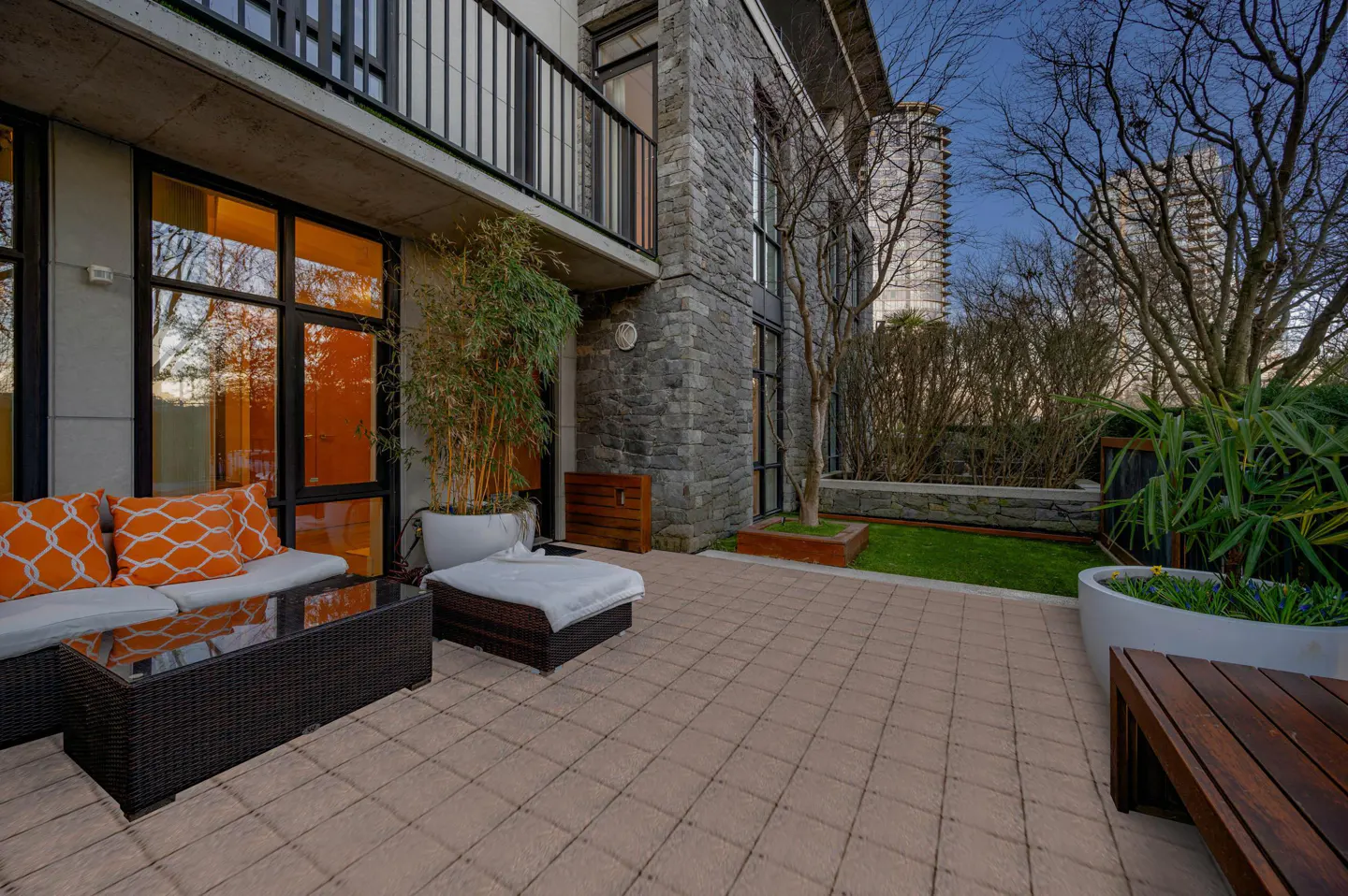 Outdoor patio with wicker furniture, orange pillows, and a glass table. A stone building and greenery are in the background.