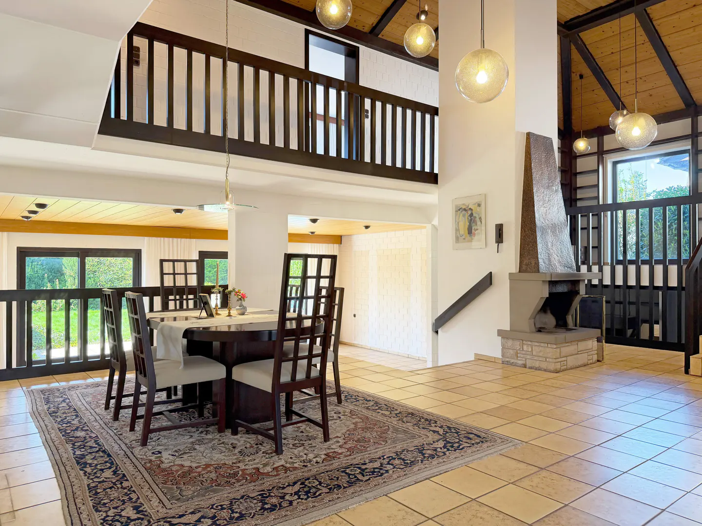 A spacious dining room with a dark wood table and chairs on a patterned rug, with a fireplace and a balcony above.