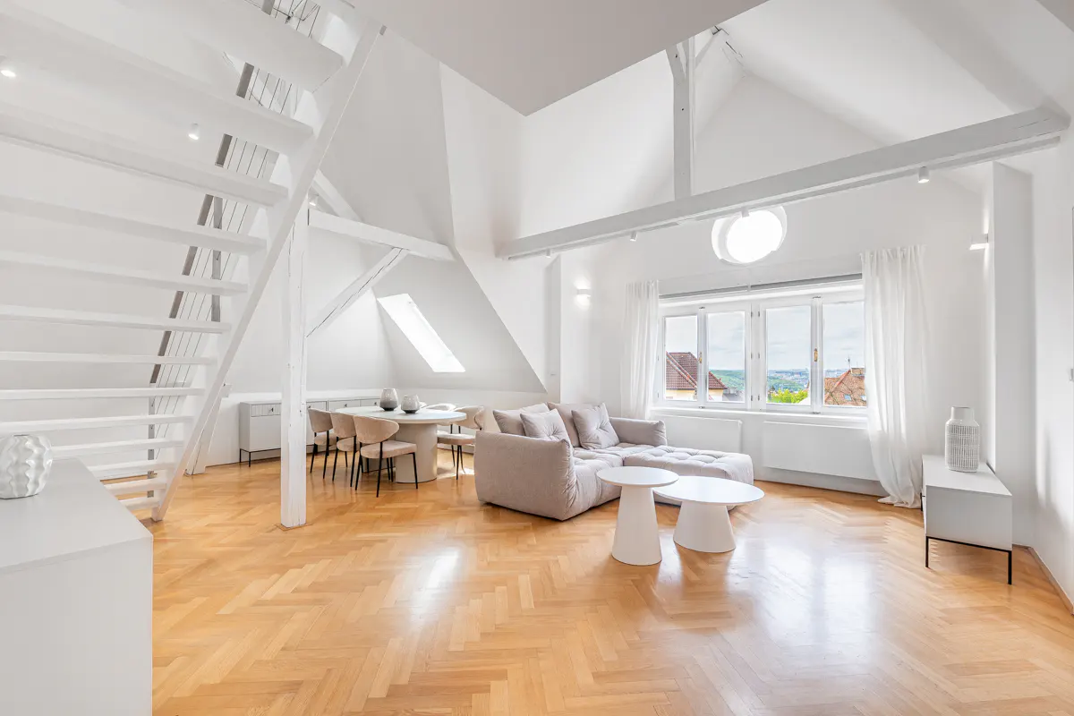 Bright, airy attic apartment with white walls, wooden floors, and exposed beams. A staircase leads up, and a large window offers a view.