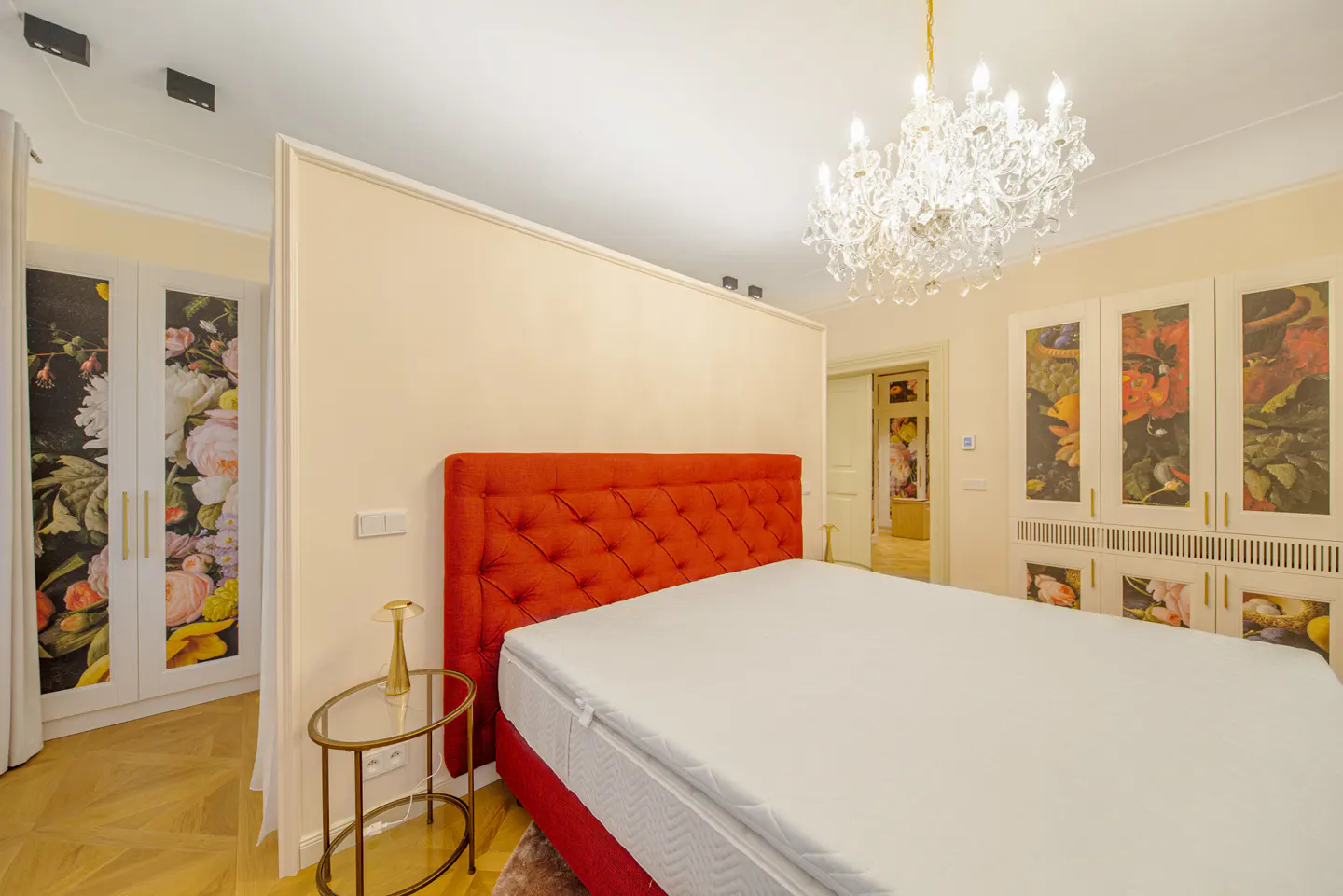 Bedroom with red tufted headboard, white bedding, crystal chandelier, floral closet doors, and gold side table.