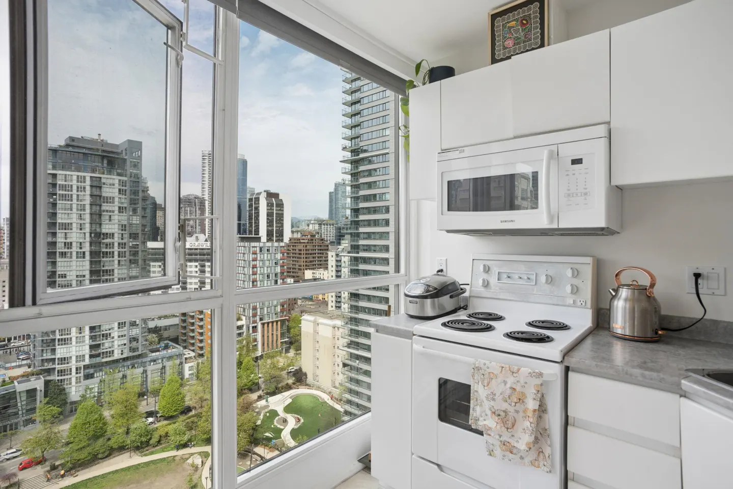 Bright kitchen with white cabinets, microwave, and stove. A window offers a city view with skyscrapers and a park.