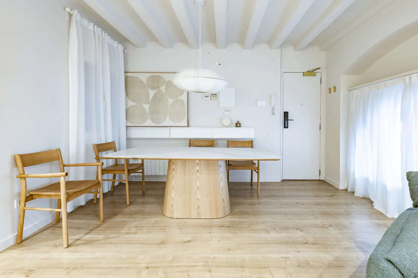 Bright, modern dining area with a white marble table, wooden chairs, and light wood floors. White walls, curtains, and ceiling beams create an airy feel.