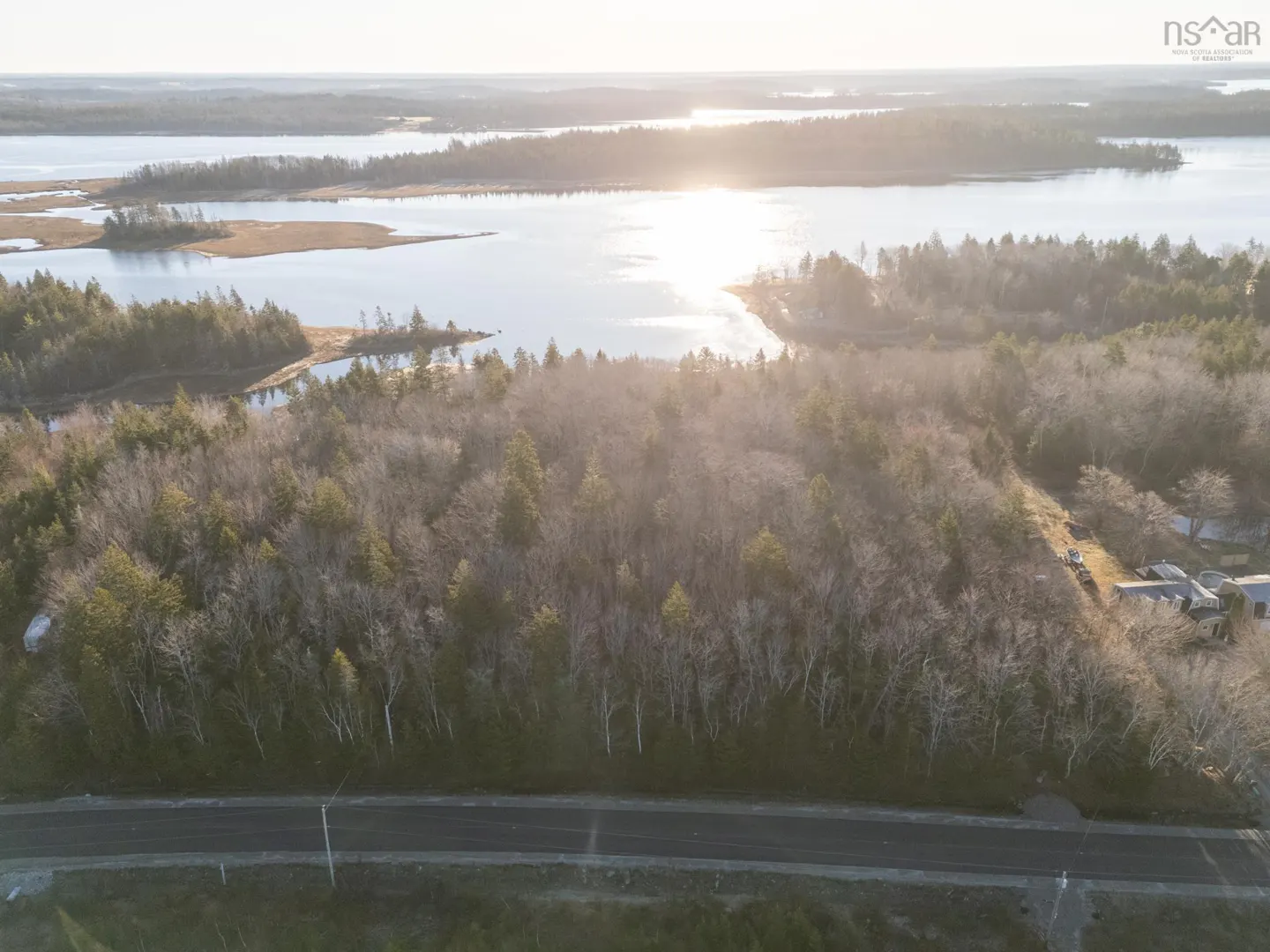 Aerial view of wooded land with a road in the foreground and a body of water in the background.