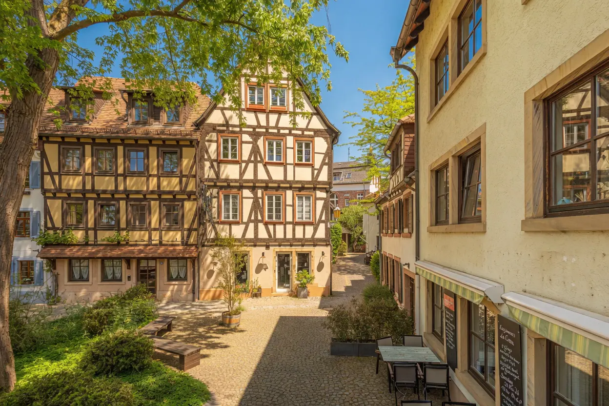 Courtyard view of half-timbered buildings with yellow walls and brown wood frames, cobblestone ground, and green foliage.