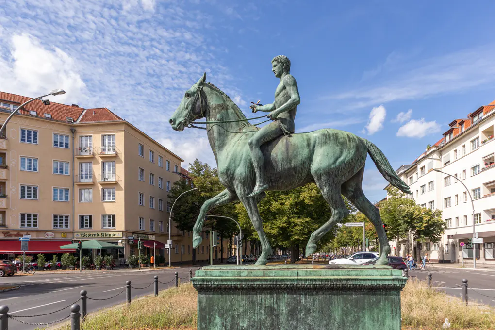 Bronze statue of a nude man riding a horse, set on a plinth. Buildings and trees in the background under a blue sky.