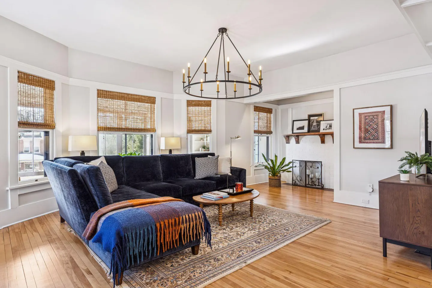 Living room with a blue sectional sofa, wood floors, and a round chandelier. Woven shades cover the windows. A fireplace is visible in the background.
