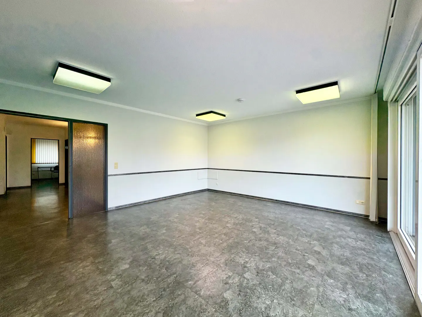 Empty room with gray floor, white walls, and three ceiling lights. A doorway leads to another room with a desk and blinds. Sliding glass doors on the right.