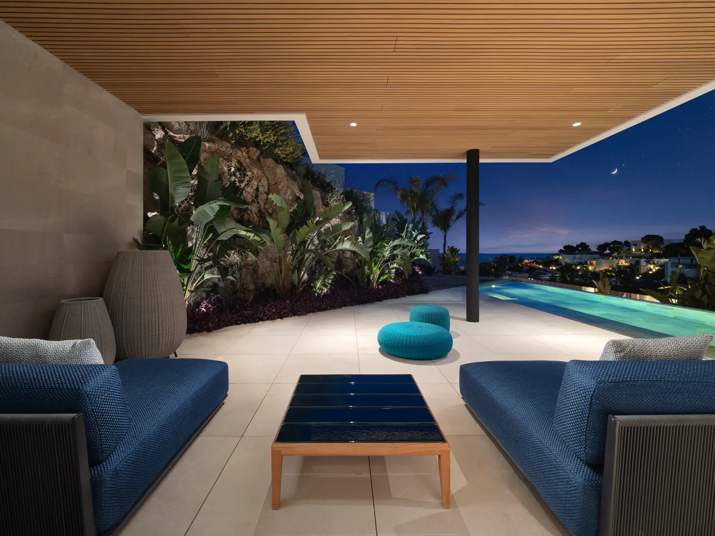 Luxury patio with blue sofas, a dark blue tiled table, and turquoise ottomans. A pool and night sky with a crescent moon are in the background.