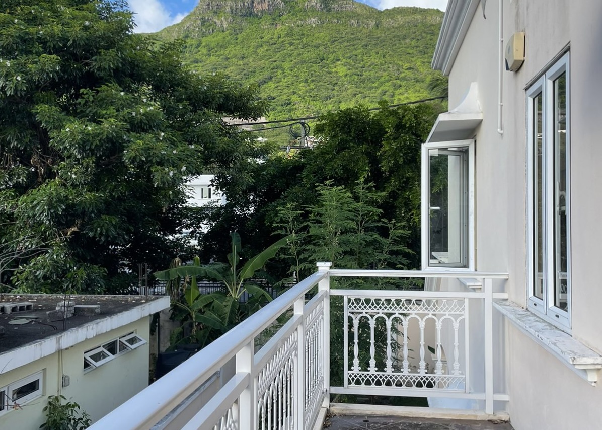 White balcony with ornate railing overlooking lush green trees and a mountain under a blue sky. [AI-generated text]