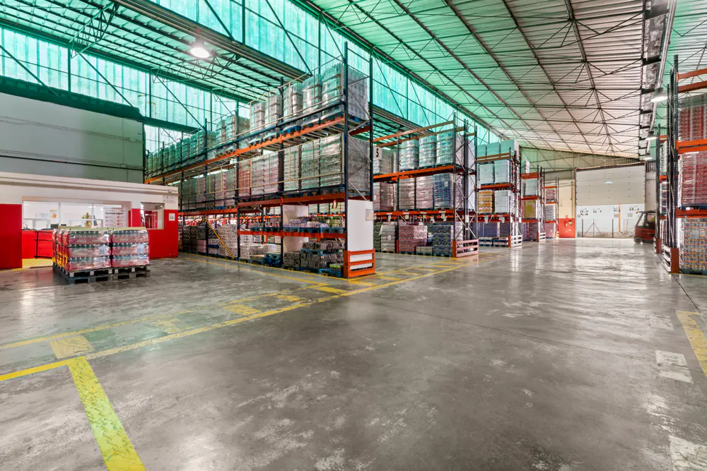 A large warehouse interior with tall shelves stocked with goods. Concrete floor with yellow lines. A red office area is visible.