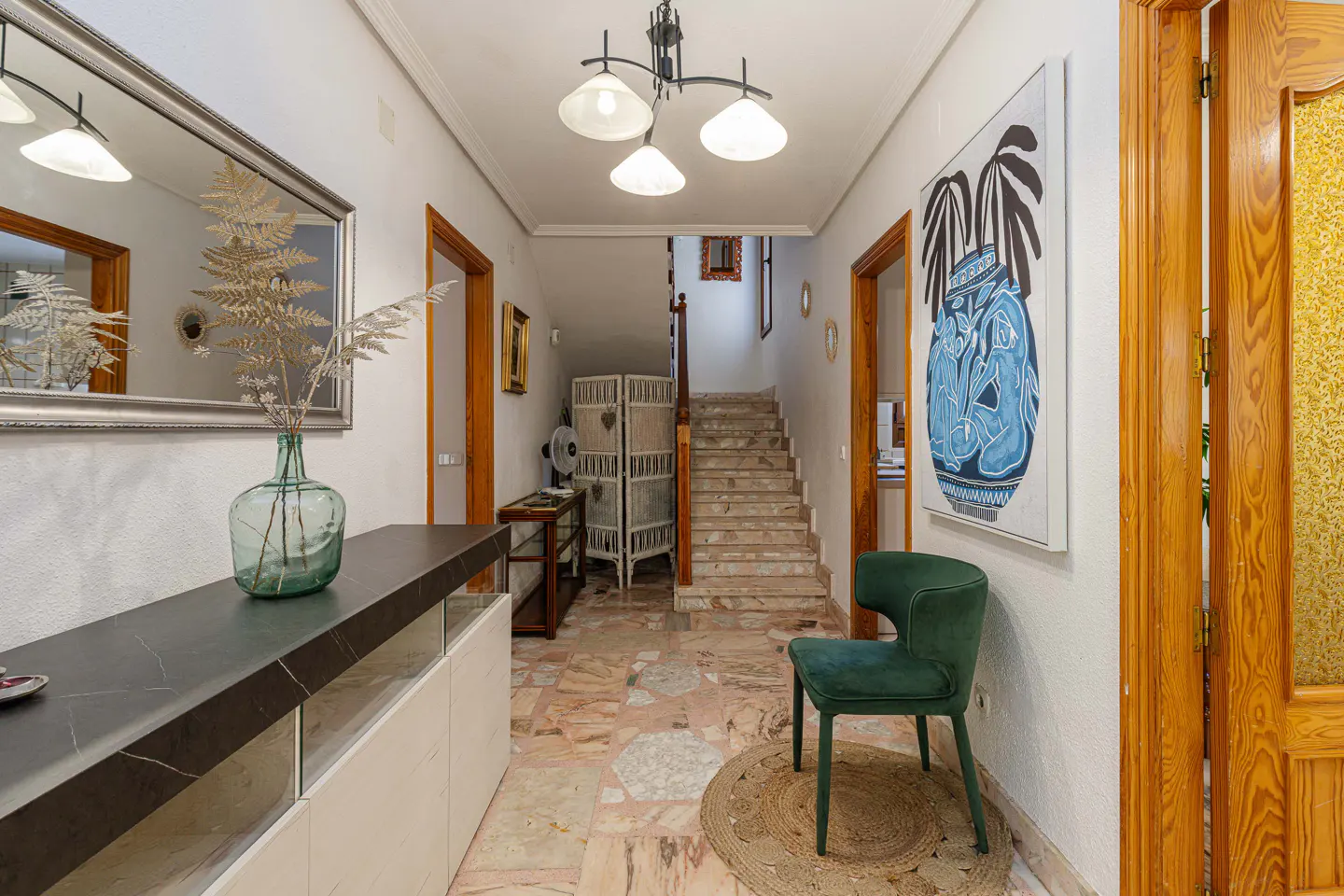Hallway with marble floors, white walls, and wooden doors. A green chair sits on a round rug. A console table with a mirror and vase is on the left. Stairs lead to the upper floor.