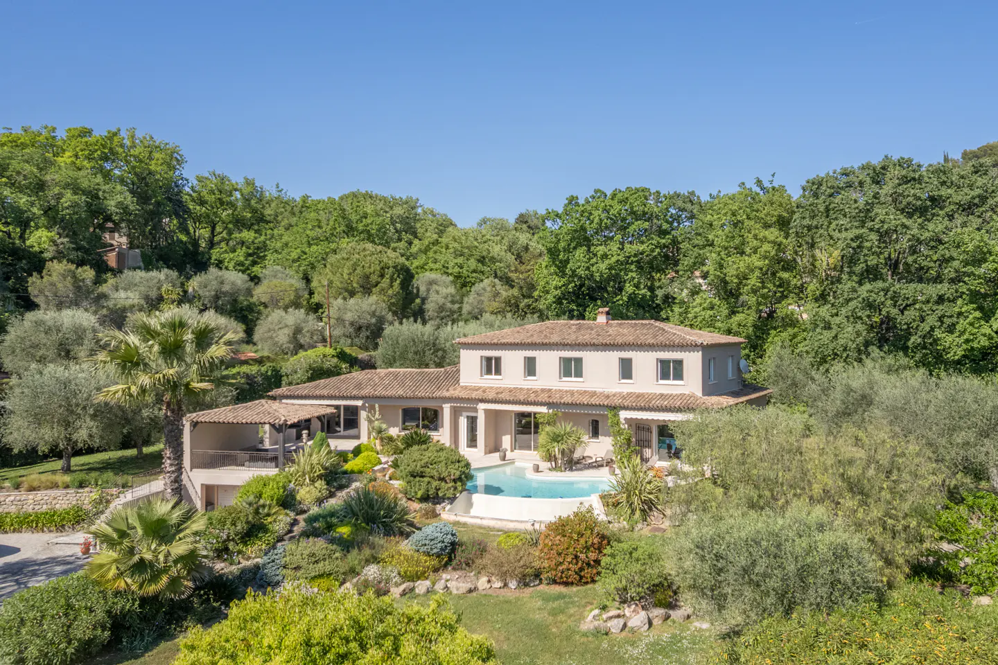 Aerial view of a two-story beige house with a red tile roof, surrounded by lush green trees and a blue swimming pool.