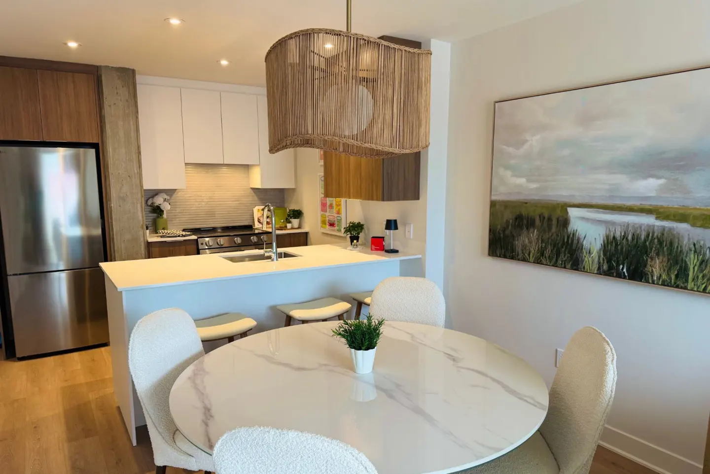 Bright, modern kitchen with a marble table, white chairs, and a woven pendant light. Stainless steel fridge and landscape art.