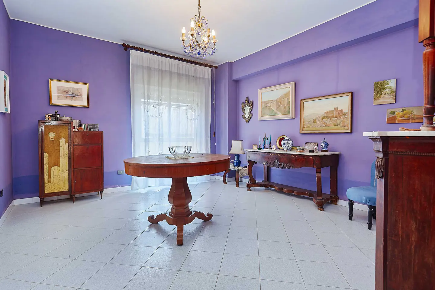 A room with purple walls, white tile floors, and antique furniture. A round wooden table sits in the center, with paintings on the walls.