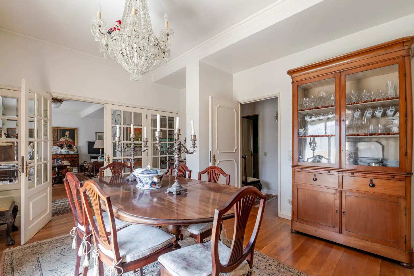 A dining room with a wood table, chairs, chandelier, and china cabinet. The walls are white and the floor is wood.