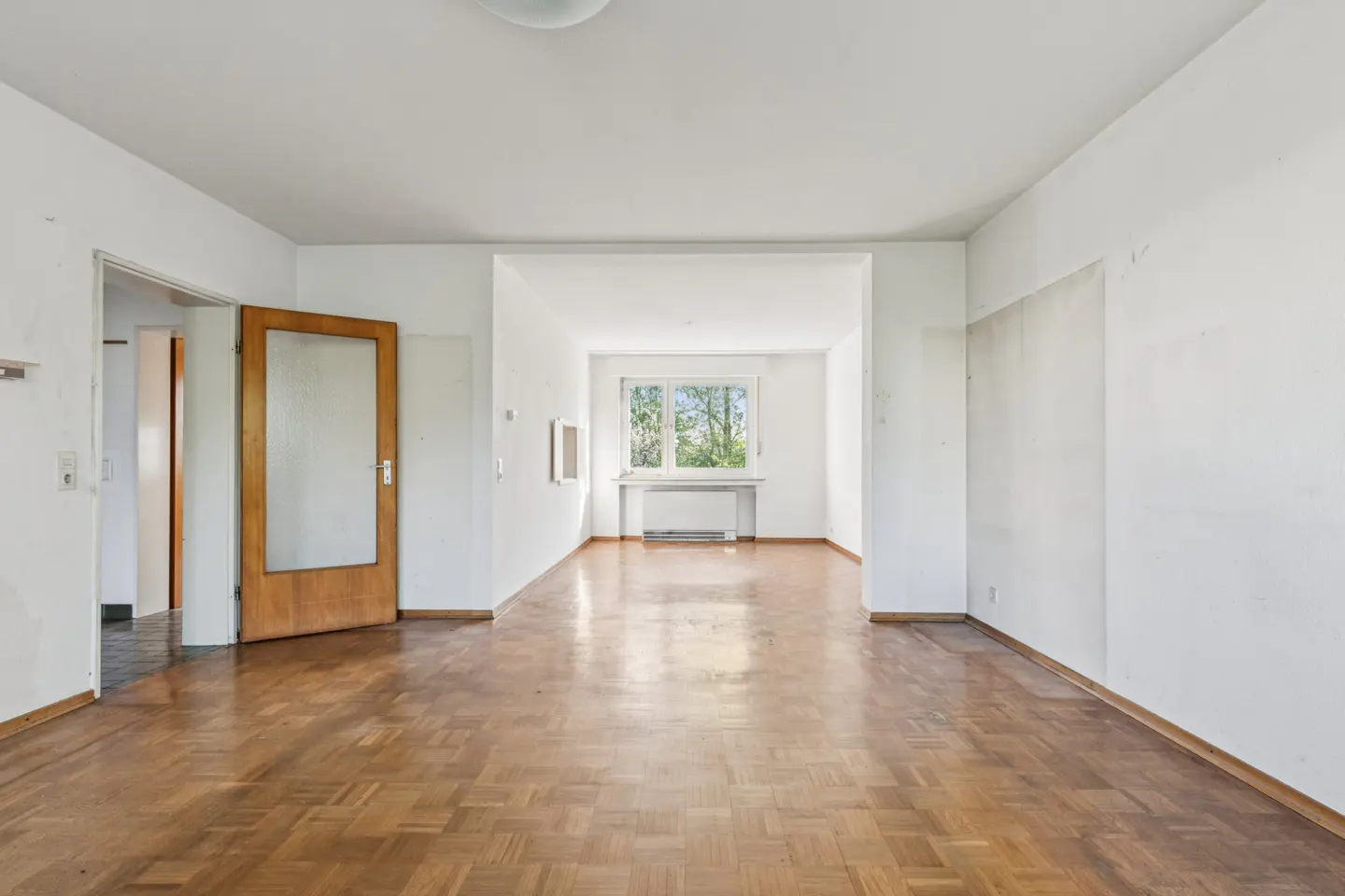 Bright, empty room with parquet floors, white walls, and a window. A wooden door with glass is slightly ajar.