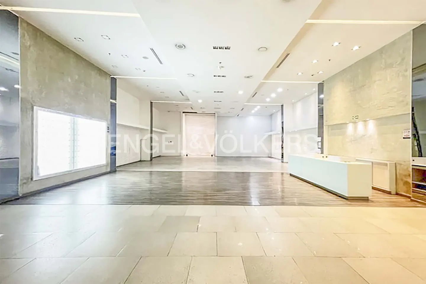 Bright, empty retail space with tile floors, white walls, and recessed lighting. A white reception desk sits on the right.