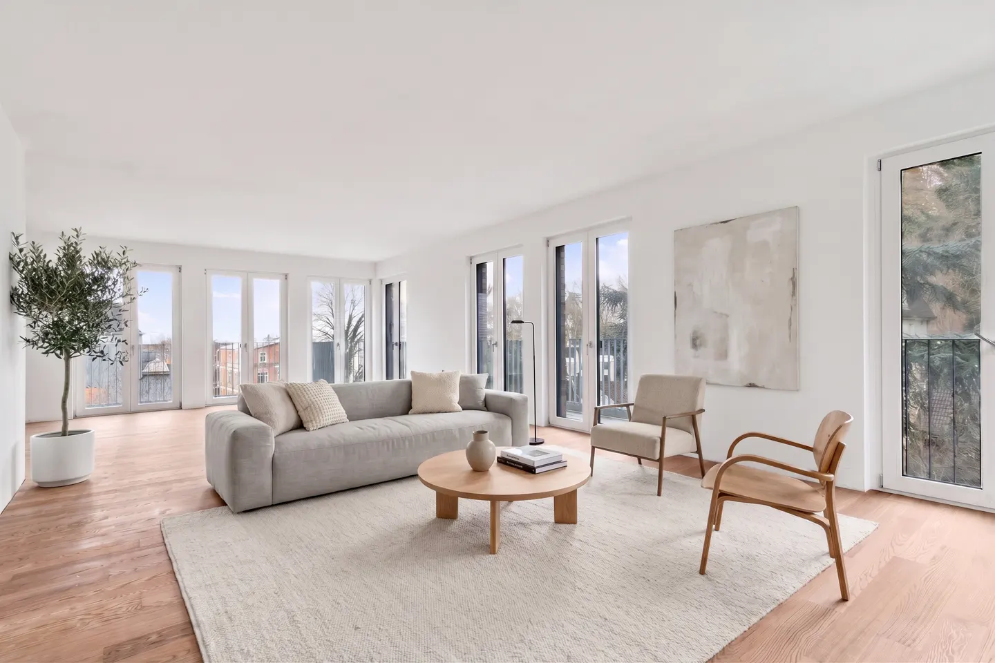 Bright living room with a gray sofa, beige chairs, and a round wooden table on a cream rug. Large windows and a potted tree add natural light.