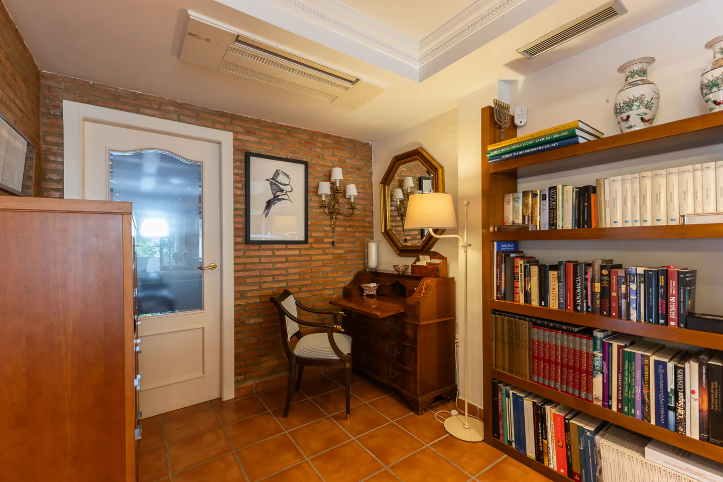 A cozy study with a brick wall, wooden desk, chair, floor lamp, and a tall bookshelf filled with many books.