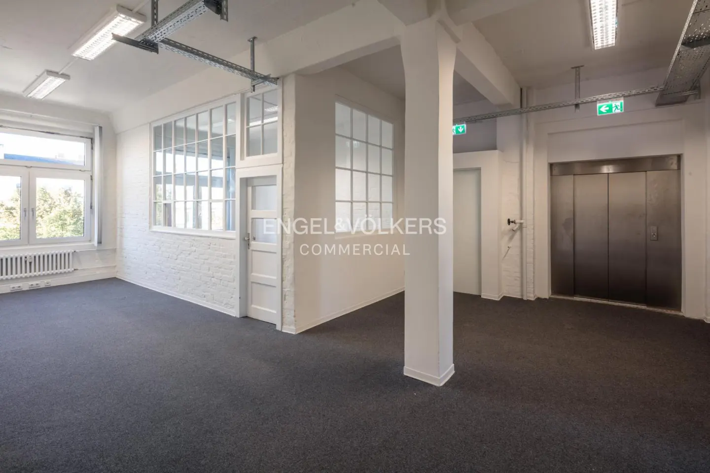 Bright, empty commercial space with gray carpet, white walls, windows, and an elevator. Overhead lighting and exposed ductwork are visible.